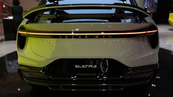 The rear view of an electric vehicle displaying a streamlined design with a prominent spoiler and a continuous LED taillight. The branding 'LOTUS' and 'ELETRE' is visible. The setting appears to be indoors, possibly an auto show or showroom.