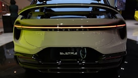 The rear view of an electric vehicle displaying a streamlined design with a prominent spoiler and a continuous LED taillight. The branding 'LOTUS' and 'ELETRE' is visible. The setting appears to be indoors, possibly an auto show or showroom.