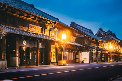 Traditional Japanese buildings with wooden facades and intricate roof designs line a quiet street. Warm streetlights cast a soft glow, highlighting the architectural details and creating a serene atmosphere. Signage with Japanese characters can be seen, enhancing the cultural essence.