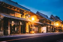 Traditional Japanese buildings with wooden facades and intricate roof designs line a quiet street. Warm streetlights cast a soft glow, highlighting the architectural details and creating a serene atmosphere. Signage with Japanese characters can be seen, enhancing the cultural essence.