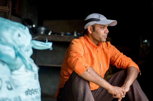 A man wearing an orange jacket and a light gray cap is sitting with his arms resting on his knees. He appears to be in deep thought or contemplation. The setting seems to be indoors, possibly in a storage area or workshop, with some utensils visible in the background and a large blue bag to his left.