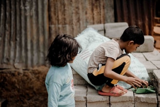 Two children are present, one standing and the other squatting on stacked bricks. The child squatting is focused on playing or working with something green in a tray, while the other child stands nearby observing. The background features corrugated metal and further stacked bricks for a rustic setting.