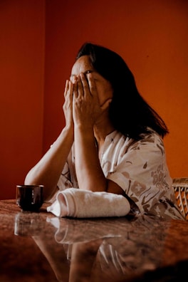 A person sits at a table with hands covering their face, conveying a sense of distress or contemplation. The table has a reflective surface with a cup and a rolled towel placed on it. The background is a warm orange color, adding depth to the scene.