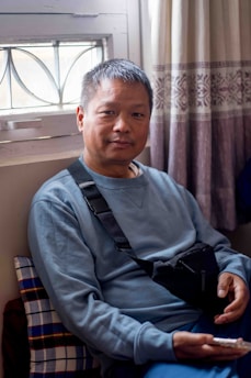 A person sitting indoors near a window with a decorative grille. They have short hair and wear a blue sweatshirt and a crossbody bag. A patterned curtain hangs beside them, and they are holding a smartphone.