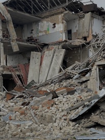 A severely damaged building with large portions of walls collapsed and debris scattered around. The structure appears to be partially demolished, with exposed beams and broken concrete slabs. Rubble consisting of bricks and twisted metal covers the ground.