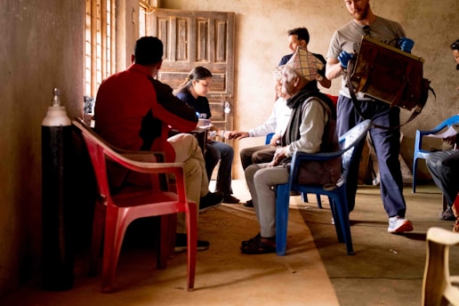 A group of people gathered in a simple, dimly lit room with sparse furniture. An older man sits on a blue plastic chair, wearing a scarf and hat, engaging with a younger woman who is writing notes. Various other individuals are seated or standing around in plastic chairs. One person carries a large box-like object. Oxygen cylinder stands against the wall.