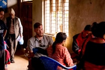 People are gathered in a dimly lit room with natural light streaming in through a window. A man wearing a stethoscope around his neck is attending to a seated woman in traditional attire. Another man stands in the background holding papers and wearing a hat. Other individuals are engaged in various activities within the room.