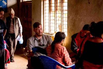 People are gathered in a dimly lit room with natural light streaming in through a window. A man wearing a stethoscope around his neck is attending to a seated woman in traditional attire. Another man stands in the background holding papers and wearing a hat. Other individuals are engaged in various activities within the room.