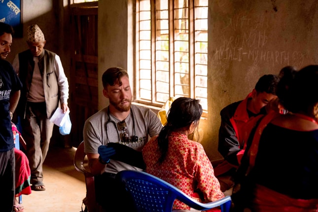 People are gathered in a dimly lit room with natural light streaming in through a window. A man wearing a stethoscope around his neck is attending to a seated woman in traditional attire. Another man stands in the background holding papers and wearing a hat. Other individuals are engaged in various activities within the room.