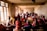 A group of people are seated in a dimly lit room, some wearing traditional clothing. They are engaging in conversation or listening intently. The room has rough, unpainted walls and is illuminated by natural light through barred windows.