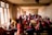 A group of people are seated in a dimly lit room, some wearing traditional clothing. They are engaging in conversation or listening intently. The room has rough, unpainted walls and is illuminated by natural light through barred windows.