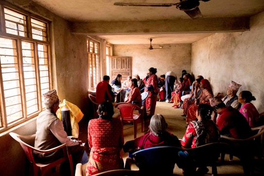 A group of people are seated in a dimly lit room, some wearing traditional clothing. They are engaging in conversation or listening intently. The room has rough, unpainted walls and is illuminated by natural light through barred windows.