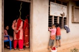 A group of people are gathered in front of a building. On the left side, three women in red and pink traditional attire stand outside an open door, one seated on a blue chair. Two children are seen on the right, peering into a window, dressed in casual clothes. Above them, a sign written in a language using a non-Latin script is posted.