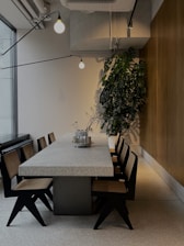 A modern dining area features a long stone table surrounded by stylish wooden chairs with woven seats. Industrial lighting hangs from the ceiling, adding an elegant touch. A tall green plant in the corner adds a natural element to the space. The walls are a combination of wood paneling and light-colored textured surfaces, contributing to a contemporary and clean ambiance.