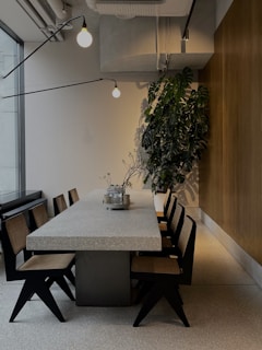 A modern dining area features a long stone table surrounded by stylish wooden chairs with woven seats. Industrial lighting hangs from the ceiling, adding an elegant touch. A tall green plant in the corner adds a natural element to the space. The walls are a combination of wood paneling and light-colored textured surfaces, contributing to a contemporary and clean ambiance.