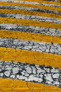 Yellow and white stripes intersect, creating a weathered asphalt texture with cracks. The alternating pattern adds an artistic element to the rough surface.