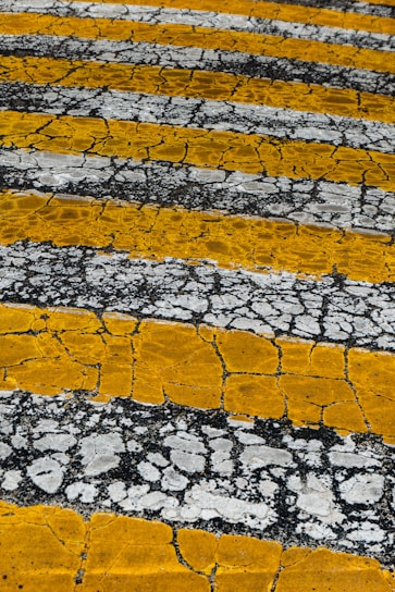 Yellow and white stripes intersect, creating a weathered asphalt texture with cracks. The alternating pattern adds an artistic element to the rough surface.