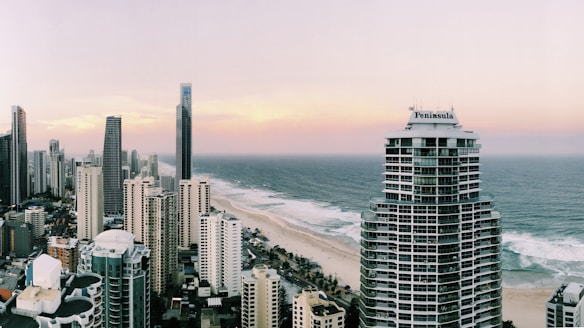 A coastal cityscape with numerous high-rise buildings lining a sandy beach. The ocean stretches on the right, with waves gently crashing onto the shore. The sky is painted in pastel shades of pink and purple, suggesting either early morning or late afternoon. One prominent building in the foreground is labeled 'Peninsula'.