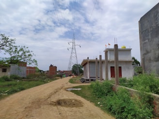 A rural setting with a dirt road stretching through the center. On either side of the road, there are small buildings with unfinished structures and tall electricity poles. In the distance, a large electricity transmission tower is visible under a partly cloudy sky. Vegetation alongside the road and near the buildings adds a touch of greenery.