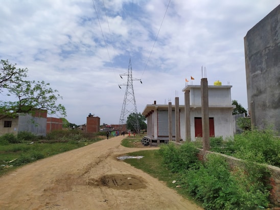 A rural setting with a dirt road stretching through the center. On either side of the road, there are small buildings with unfinished structures and tall electricity poles. In the distance, a large electricity transmission tower is visible under a partly cloudy sky. Vegetation alongside the road and near the buildings adds a touch of greenery.