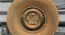 A close-up view of a heavily worn and dusty tire mounted on a metal rim, with visible bolts and text imprinted on the sidewall. The background features a vehicle's body panel in a muted color.