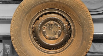 A close-up view of a heavily worn and dusty tire mounted on a metal rim, with visible bolts and text imprinted on the sidewall. The background features a vehicle's body panel in a muted color.
