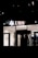 The image depicts the exterior of a UBS bank branch at night, with illuminated signage displaying the UBS logo and letters. The predominantly dark surroundings contrast with the brightly lit interior visible through the large glass windows.