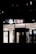 The image depicts the exterior of a UBS bank branch at night, with illuminated signage displaying the UBS logo and letters. The predominantly dark surroundings contrast with the brightly lit interior visible through the large glass windows.