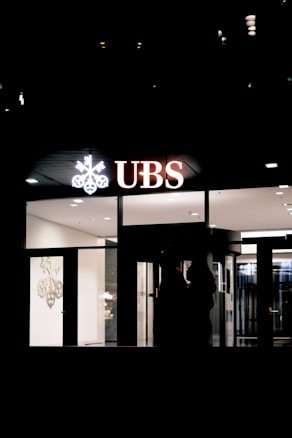 The image depicts the exterior of a UBS bank branch at night, with illuminated signage displaying the UBS logo and letters. The predominantly dark surroundings contrast with the brightly lit interior visible through the large glass windows.