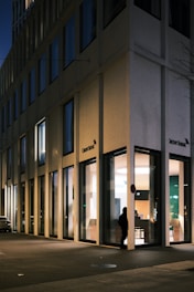 A modern office building at night with large glass windows reflecting city lights. A silhouette of a person stands near the entrance, surrounded by soft interior lighting that contrasts with the dim exterior.