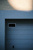 A garage door with horizontal panels and a small rectangular window located near the top left corner. Above the door, there's a section of textured brick wall illuminated by light, creating a contrast between the bright and shadowed areas.