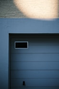 A garage door with horizontal panels and a small rectangular window located near the top left corner. Above the door, there's a section of textured brick wall illuminated by light, creating a contrast between the bright and shadowed areas.