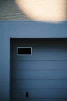 A garage door with horizontal panels and a small rectangular window located near the top left corner. Above the door, there's a section of textured brick wall illuminated by light, creating a contrast between the bright and shadowed areas.