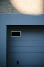 A garage door with horizontal panels and a small rectangular window located near the top left corner. Above the door, there's a section of textured brick wall illuminated by light, creating a contrast between the bright and shadowed areas.