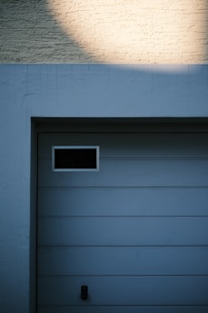A garage door with horizontal panels and a small rectangular window located near the top left corner. Above the door, there's a section of textured brick wall illuminated by light, creating a contrast between the bright and shadowed areas.