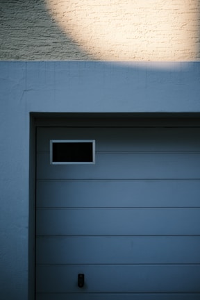 A garage door with horizontal panels and a small rectangular window located near the top left corner. Above the door, there's a section of textured brick wall illuminated by light, creating a contrast between the bright and shadowed areas.