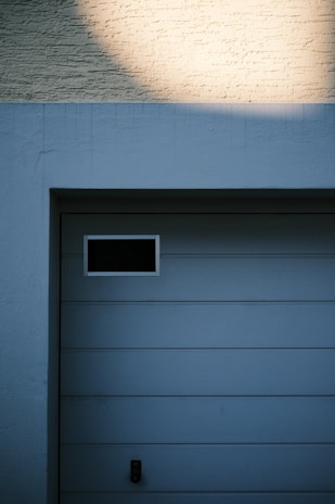 A garage door with horizontal panels and a small rectangular window located near the top left corner. Above the door, there's a section of textured brick wall illuminated by light, creating a contrast between the bright and shadowed areas.