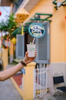 A hand holding a paper cup with a straw and a leaf garnish in front of a yellow building with a green and blue sign. The scene includes colorful lanterns and a small gate.