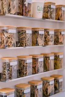 Multiple shelves hold square glass jars with bamboo lids, each containing various dried herbs and leaves neatly organized. Labels are attached to each jar. A white paper cup with green text is placed among the jars.