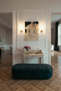 A sophisticated, elegant room with classic decor. On the wall, there is a painting of a woman in a white dress, flanked by two sconces emitting soft, warm light. Below the painting, a table holds a vase with pink flowers, a small gold tray, and a candle. A plush, dark green bench sits in front of the table. The room features parquet flooring and large windows with white curtains, allowing natural light to illuminate the space.