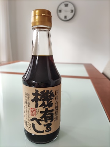 A bottle of soy sauce with Japanese text on the label sits on a reflective, glass-topped table. The bottle has a gold cap and a tan-colored label featuring bold black characters. In the blurred background, a wall clock is visible.