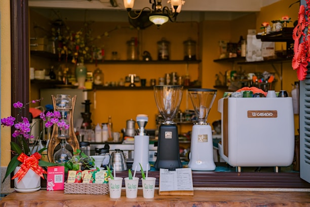A cozy coffee shop counter displaying two coffee grinders, a coffee machine, and a variety of utensils. There are small pots with flowers, a red ribbon wraps around a basket of items, and a few takeaway cups labeled 'Cà Lao' in the foreground. Shelves in the background hold various jars and decorative items, contributing to a warm and inviting atmosphere.