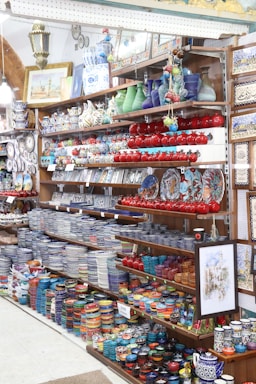 The image features a well-stocked store shelf filled with a variety of colorful ceramic and pottery items. There are numerous bowls, plates, and vases, most displaying intricate patterns and vibrant colors such as blue, red, and green. Decorative lanterns and framed artwork are also visible, along with small ceramic fruit ornaments.