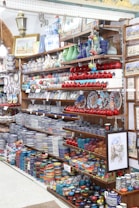 The image features a well-stocked store shelf filled with a variety of colorful ceramic and pottery items. There are numerous bowls, plates, and vases, most displaying intricate patterns and vibrant colors such as blue, red, and green. Decorative lanterns and framed artwork are also visible, along with small ceramic fruit ornaments.