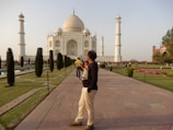 A man stands on a paved walkway in front of the Taj Mahal, which is visible in the background with its iconic white marble dome and four minarets. The area is busy with tourists walking around the manicured gardens featuring pathways and small bushes. The sky is clear and the scene is well-lit by daylight.