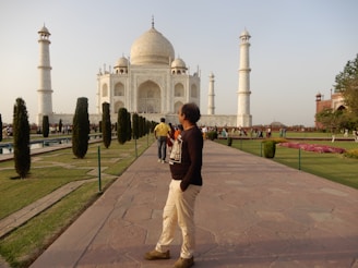 A man stands on a paved walkway in front of the Taj Mahal, which is visible in the background with its iconic white marble dome and four minarets. The area is busy with tourists walking around the manicured gardens featuring pathways and small bushes. The sky is clear and the scene is well-lit by daylight.