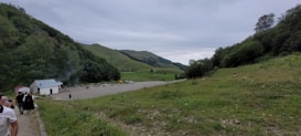 A serene landscape with a small building and a group of people on a paved area. Rolling green hills and a lush forest are visible under a cloudy sky. A few parked vehicles are in the vicinity, and scattered chairs suggest a gathering or resting spot.
