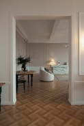A stylish living room with parquet wooden flooring, featuring a round wooden table with a black vase containing branches and a chair. A contemporary white armchair is positioned nearby, facing a modern fireplace with a marble finish. An elegant table lamp on a sideboard provides soft lighting, and a framed picture adds a decorative touch.