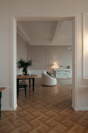 A stylish living room with parquet wooden flooring, featuring a round wooden table with a black vase containing branches and a chair. A contemporary white armchair is positioned nearby, facing a modern fireplace with a marble finish. An elegant table lamp on a sideboard provides soft lighting, and a framed picture adds a decorative touch.
