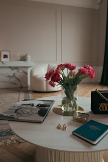 A chic, elegantly furnished living room with a round marble coffee table adorned with various items such as a fashion magazine, a small notebook, a glass perfume bottle, and a couple of rings. A vase filled with vibrant pink tulips adds a pop of color to the subdued color scheme. In the background, there is a plush white armchair and a white marble console table with decorative items.